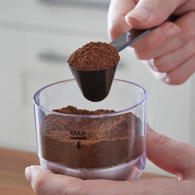 Person holding a black measuring scoop filled with finely ground coffee above a clear plastic container marked with MAX line, containing ground coffee, demonstrating the Morphy Richards Brew and Blend electric coffee grinder