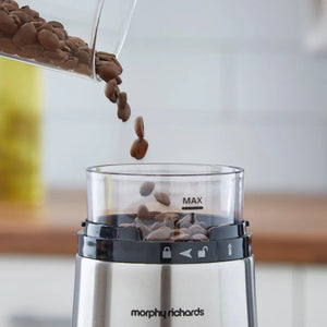 Morphy Richards Brew and Blend electric coffee grinder in stainless steel pouring roasted coffee beans into the grinding chamber, showing the MAX fill line and control buttons on the black band