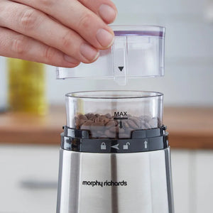 Hand holding transparent water reservoir above Morphy Richards stainless steel electric coffee grinder with black control band and coffee beans visible in grinding chamber marked with MAX line