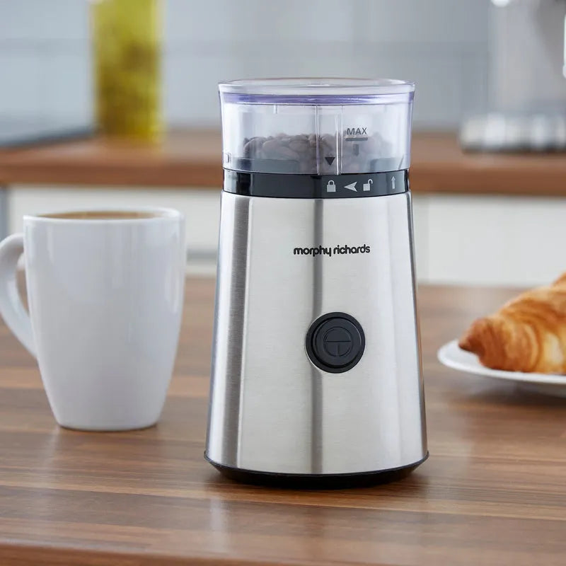 Morphy Richards stainless steel electric coffee grinder on wooden countertop with white mug of coffee and croissant, featuring transparent grinding chamber with black control band and power button