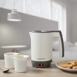 Morphy Richards compact white and grey travel kettle 0.5L on wooden countertop with two white mugs of tea and plate of biscuits in bright kitchen setting