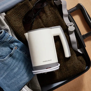 Morphy Richards white and grey compact travel kettle with 0.5L capacity displayed in a black carrying bag alongside sunglasses, jeans, and other travel items on a wooden surface