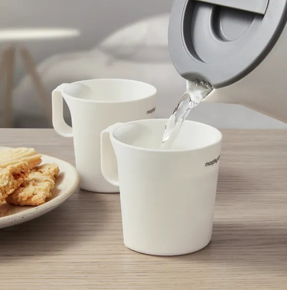 Morphy Richards white compact travel kettle pouring hot water into matching white mug on wooden table with plate of biscuits and second mug in background