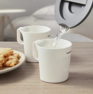 Morphy Richards white compact travel kettle pouring hot water into matching white mug on wooden table with plate of biscuits and second mug in background