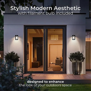 Modern outdoor entryway with black framed glass doors and white walls featuring two black solar LED wall lanterns with filament bulbs flanking the entrance, complemented by potted topiary plants and warm ambient lighting