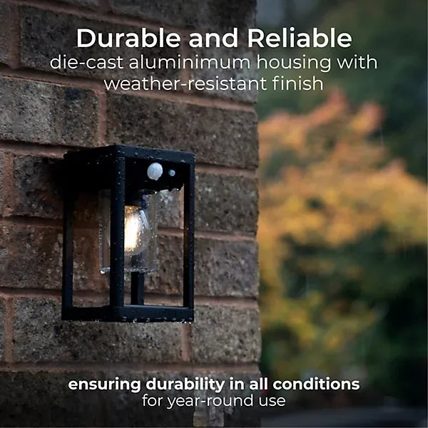 Black die-cast aluminium outdoor solar wall lantern with PIR motion sensor mounted on brick wall, featuring weather-resistant finish and illuminated LED light, with blurred garden foliage in background