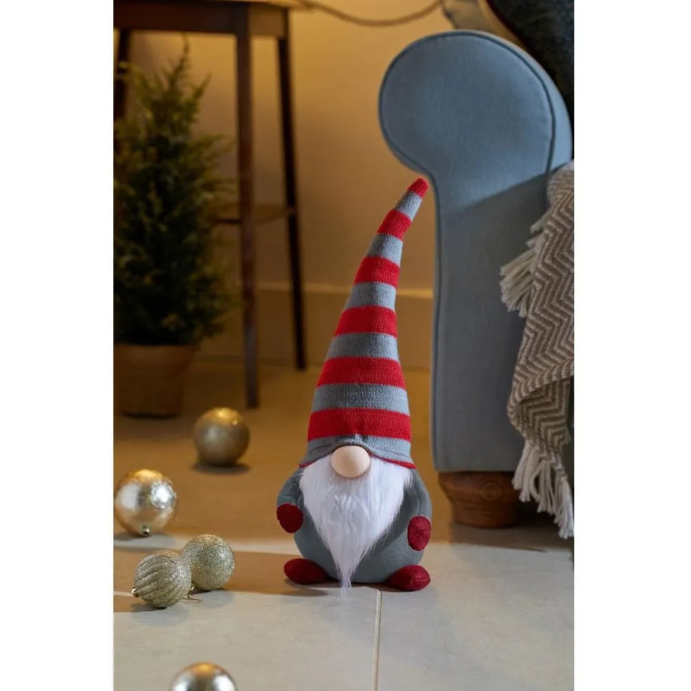 Red and grey striped knitted gnome with white beard and red pom pom details displayed on wooden floor with gold baubles and blue chair in background