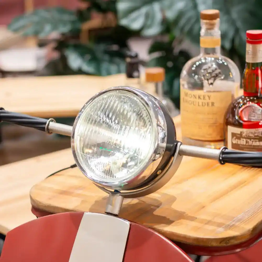 Red and white Vespa scooter-style wine rack with chrome headlight and handlebar details, displaying bottles of spirits and liqueurs on a wooden surface with potted plants in background