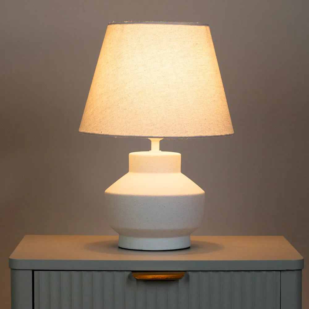 Tara Lane Chloe ceramic bedside lamp with white textured base and beige linen shade on grey cabinet, illuminated against neutral background