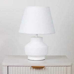 White ceramic table lamp with bulbous base and conical linen shade sitting on cream ribbed bedside cabinet against neutral background