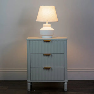 White ceramic table lamp with cream linen shade on blue-grey bedside cabinet with three drawers and brass handles