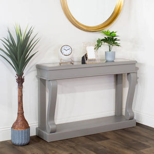 Mink grey Tara Lane Scroll Console Table with antique mirror top and curved legs, styled with potted plants, clock and decorative accessories against white wall with gold circular mirror above