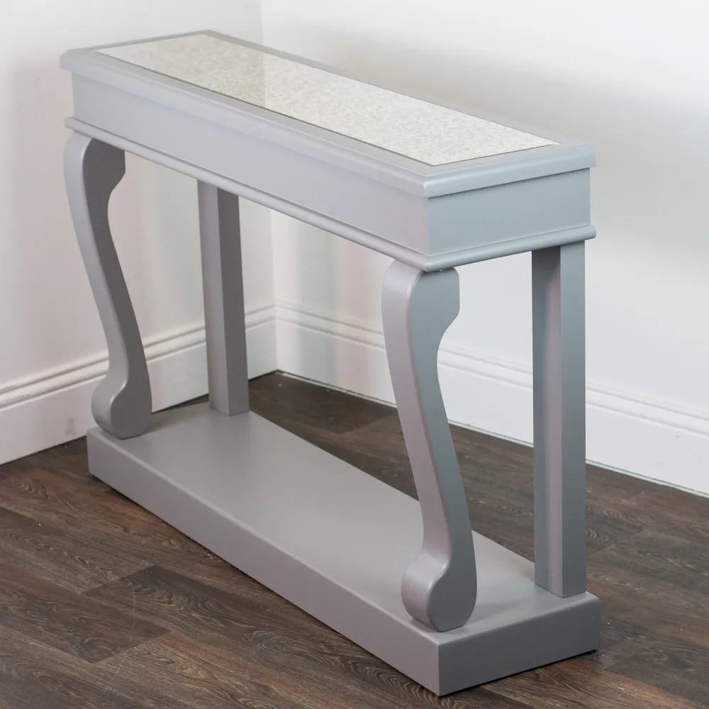 Tara Lane Scroll Console Table with mink grey finish, antique mirror top, curved bracket supports and dark wood floor placement against white wall