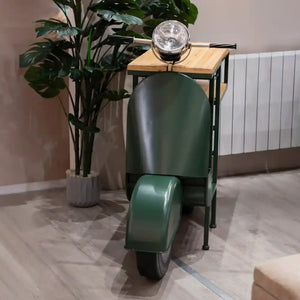 Green vintage-style Vespa scooter wine rack with natural wood top shelf, chrome headlight, black metal frame and wheel, displayed indoors next to potted plant and radiator