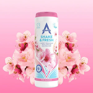 Astonish Shake and Fresh Pink Blossom carpet freshener 400g bottle with white container and pink lid, surrounded by pink and magenta flowers on pink background, showing product packaging with vegan certification logo
