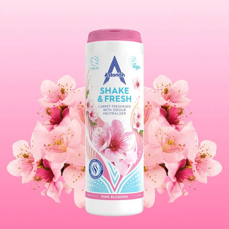 Astonish Shake and Fresh Pink Blossom carpet freshener 400g bottle with white container and pink lid, surrounded by pink and magenta flowers on pink background, showing product packaging with vegan certification logo