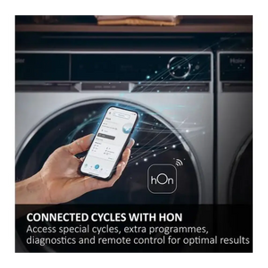 Hand holding smartphone displaying hOn app interface in front of white Haier washing machine with dark drum, showing connected cycles control feature with WiFi connectivity icon