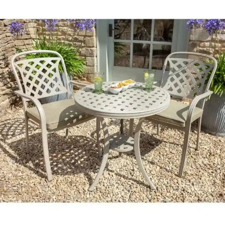 Hartman Berkeley white cast aluminium bistro set with round table and two lattice-back chairs on gravel patio, featuring cushioned seating and refreshments on table, with purple flowering plants and stone wall in background
