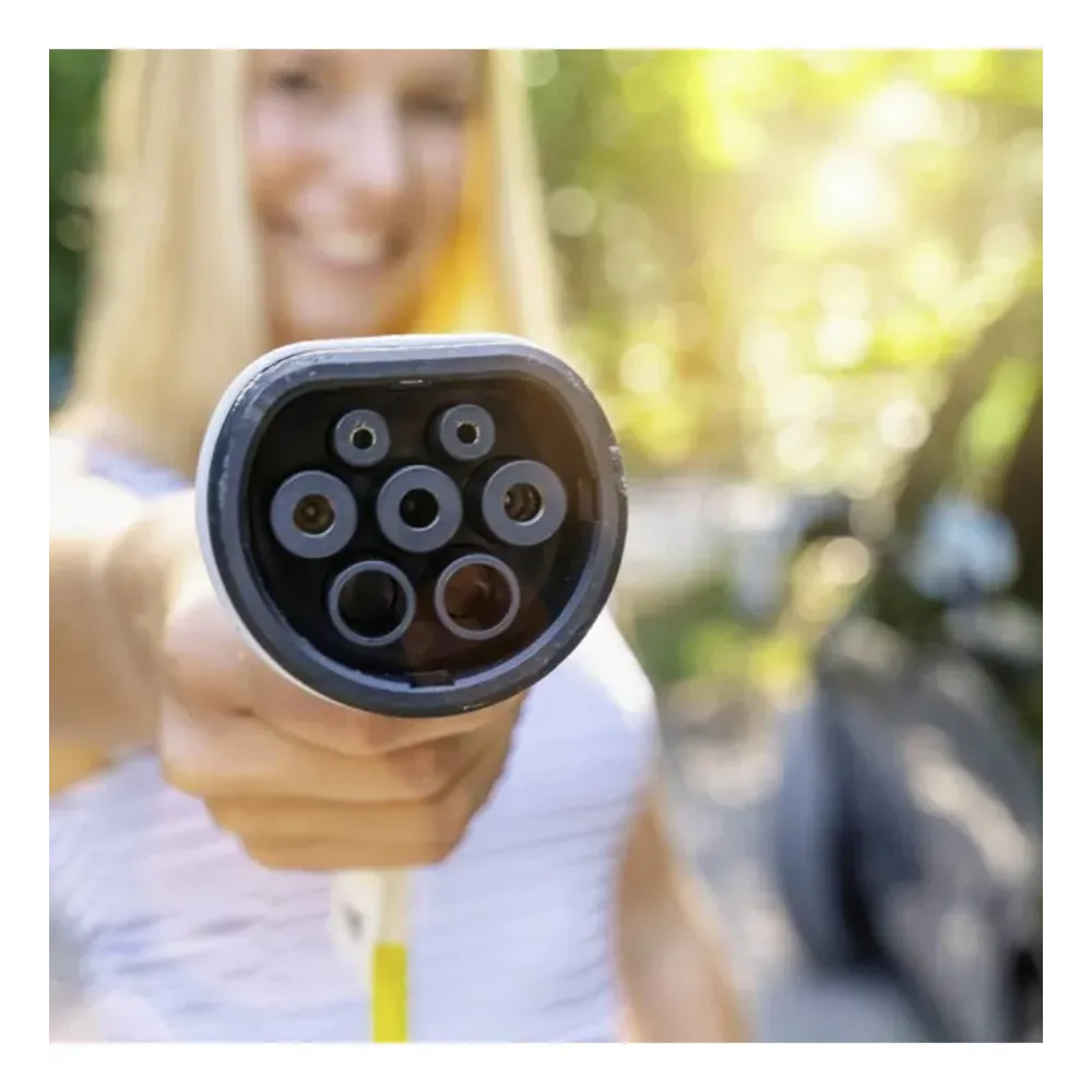 Close-up of a Type 2 EV charging connector head showing seven circular metal contact pins arranged in a circle on a black background, held by a person in a white shirt with blurred green garden setting behind