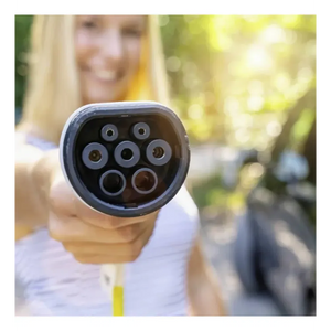 Close-up of a Type 2 EV charging connector head showing seven circular metal contact pins arranged in a circle on a black background, held by a person in a white shirt with blurred green garden setting behind