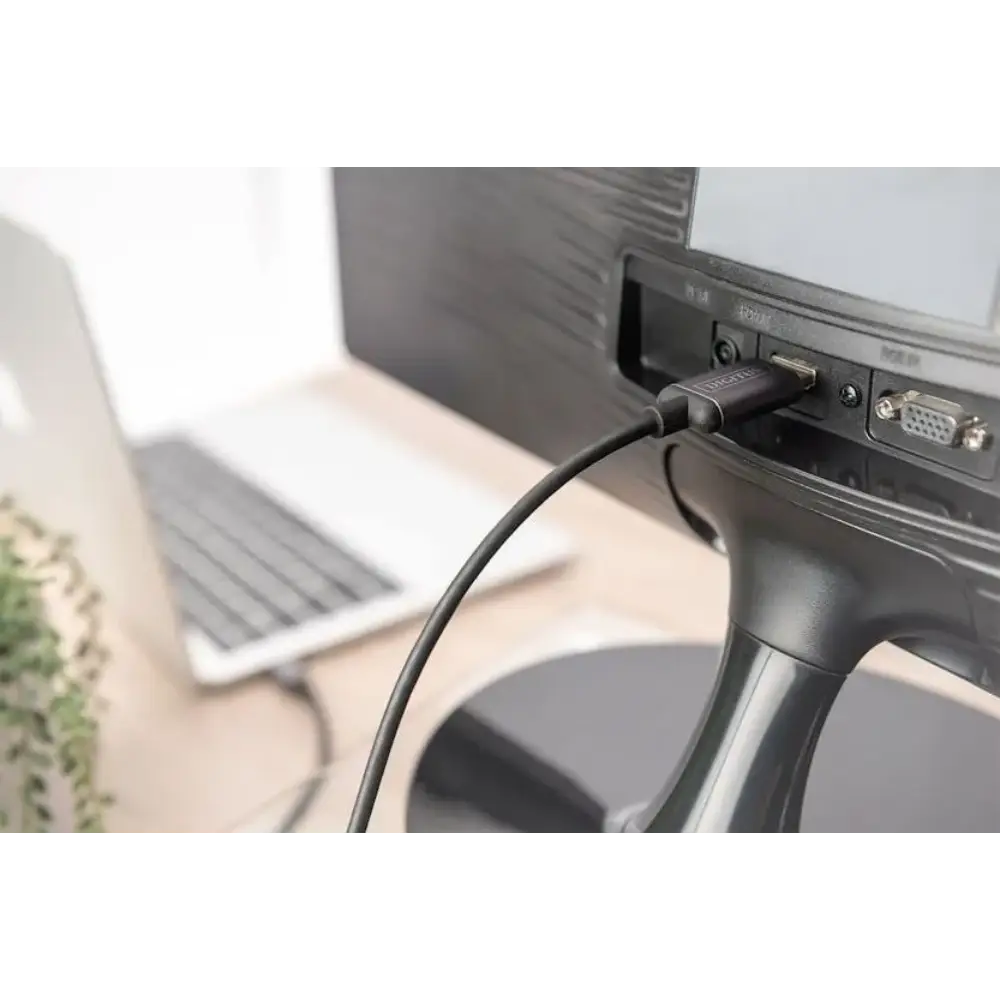Black USB-C to HDMI adapter cable connected to a monitor with VGA port in an office workspace with laptop and potted plant visible in background