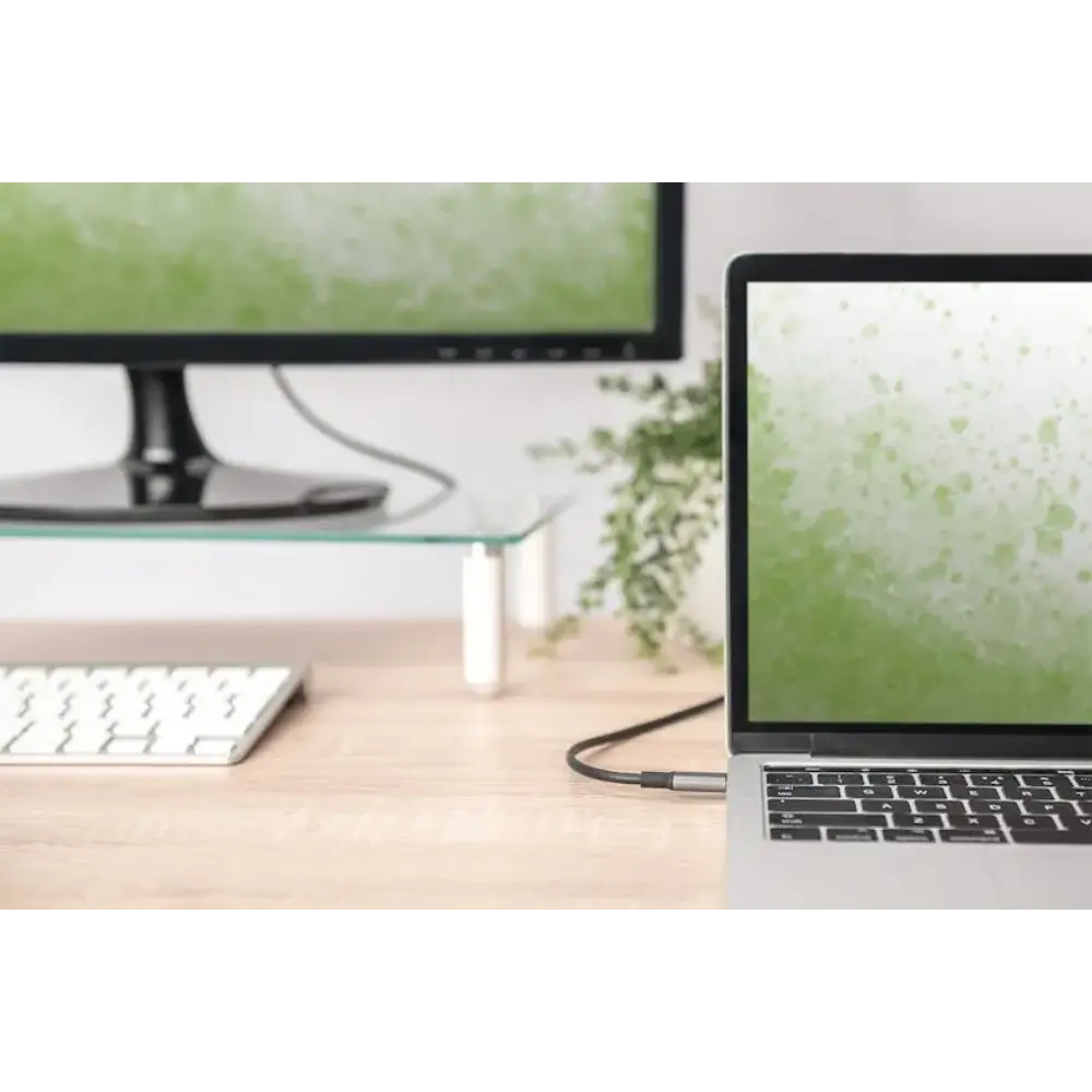 USB-C to HDMI adapter cable connecting a MacBook laptop to an external monitor in a modern office workspace with wooden desk, wireless keyboard, and potted plant