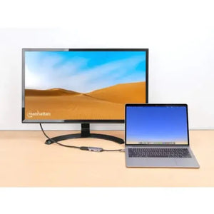 Manhattan USB C to HDMI 3 in 1 docking converter with power delivery shown connected between a silver laptop and large external monitor displaying a desert landscape on a wooden desk