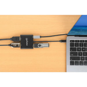 Manhattan USB-C to Dual HDMI multiport adapter with 100W power delivery connected to MacBook Pro on wooden desk, showing black adapter hub with multiple cables and USB flash drive