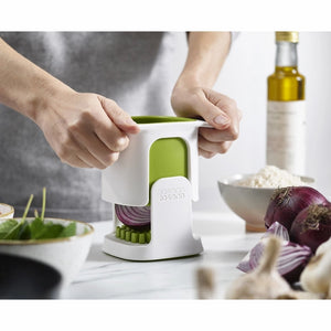 Person using white Joseph Joseph ChopCup vegetable dicer with green lid to chop red onion, with olive oil bottle and fresh ingredients visible on white kitchen counter
