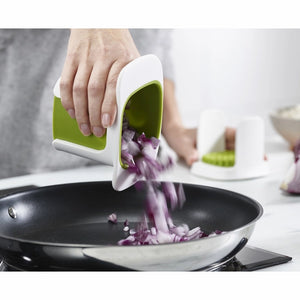 Hand using a white and green Joseph Joseph ChopCup vegetable dicer to chop purple onions into a black non-stick frying pan in a modern kitchen setting