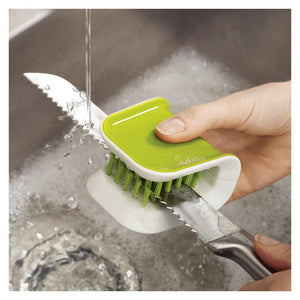 Joseph Joseph BladeBrush Knife Cleaner in green and white being used to clean a serrated knife under running water at a stainless steel sink