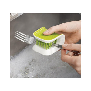 Hand holding a green and white Joseph Joseph BladeBrush knife cleaner under running water, cleaning the tines of a metal fork with built-in brush bristles