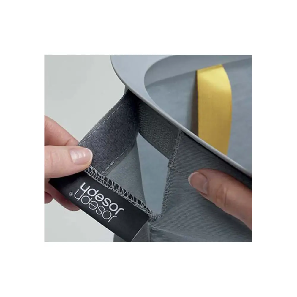 Close-up of Joseph Joseph Tota 60 litre grey laundry basket showing black fabric divider with brand label and yellow adhesive tape strip on grey compartment lid