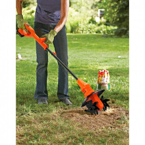 Man using Black and Decker BETL1820L cordless garden tiller with orange and black housing to cultivate soil in residential lawn, showing front tine cultivation head in action on garden bed