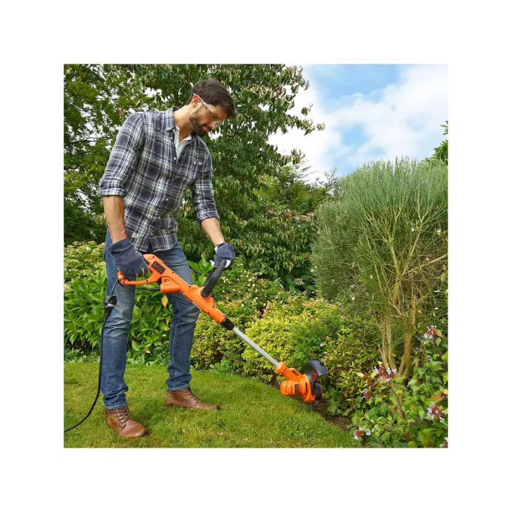 Man wearing checkered shirt and gloves using Black & Decker orange and black electric grass strimmer to trim weeds along garden flower bed in sunny garden setting