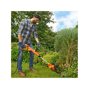 Man wearing checkered shirt and gloves using Black & Decker orange and black electric grass strimmer to trim weeds along garden flower bed in sunny garden setting