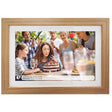 Light oak wooden digital photo frame displaying a birthday party scene with a girl blowing out candles on a decorated cake surrounded by family and friends, with white mat border and social media post caption visible at bottom