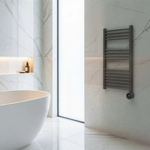 Modern grey heated towel rail mounted on marble-effect bathroom wall above white floor tiles next to freestanding white bathtub and illuminated alcove shelf