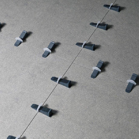 Overhead view of black and grey plastic tile levelling wedges arranged in rows on a concrete surface with white guide lines, showing the Bihui tile levelling system components