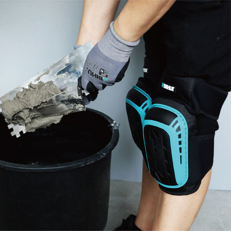 Worker wearing gray socks and black knee pads with turquoise TPR padding scooping wet cement into a black bucket with gloved hands