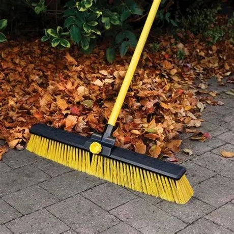 Bulldozer 24 inch heavy duty broom with yellow bristles and black brush head attached to yellow handle, positioned on pavement with autumn leaves in background