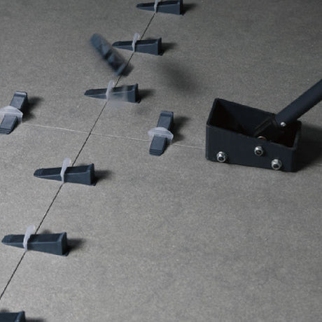 Black and gray plastic wedge leveling tile clips arranged in a line on concrete surface with adjustment tool and metal mounting bracket demonstrating proper tile spacing installation