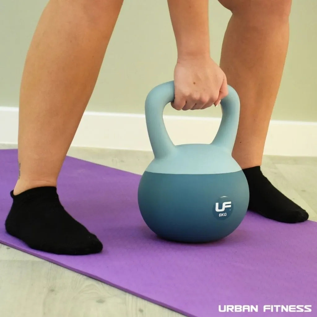 Person holding a blue and teal Urban Fitness 6kg dual soft kettlebell on a purple exercise mat, wearing black socks, demonstrating proper kettlebell grip for fitness training