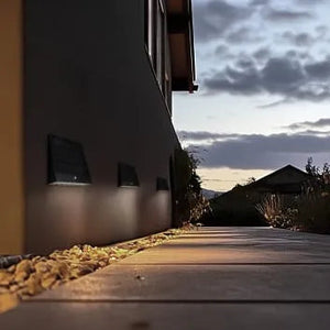 Luceco Stealth black solar wall light mounted on modern white rendered building exterior at dusk, illuminating gravel pathway with warm LED glow against twilight sky