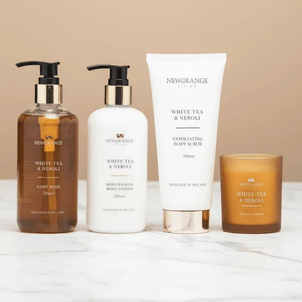 Newgrange Living White Tea and Neroli four-piece body care set displaying a brown glass hand wash bottle, white moisturising body lotion bottle, white exfoliating body scrub tube, and amber glass scented candle arranged on a marble surface against a beige background