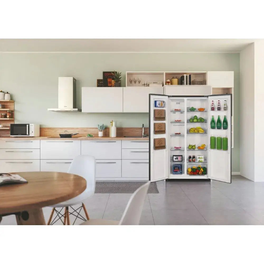 Candy 442L frost-free American fridge freezer in stainless steel displayed open in a modern kitchen with white cabinetry, wooden countertops, and mint green walls, showing well-stocked shelves with fresh produce, beverages, and groceries