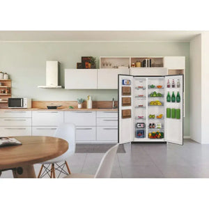Candy 442L frost-free American fridge freezer in stainless steel displayed open in a modern kitchen with white cabinetry, wooden countertops, and mint green walls, showing well-stocked shelves with fresh produce, beverages, and groceries