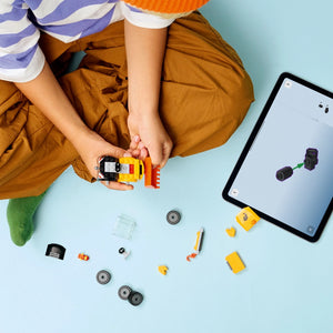Child wearing orange beanie and brown jacket holding yellow and black LEGO construction loader brick set with assembled vehicle, while tablet displays building instructions, surrounded by loose LEGO pieces and metal axles on light blue background