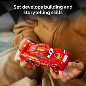 Child's hands holding a red LEGO Lightning McQueen car model numbered 95 with yellow and white details, wearing a tan brown jacket, with text above stating Set develops building and storytelling skills