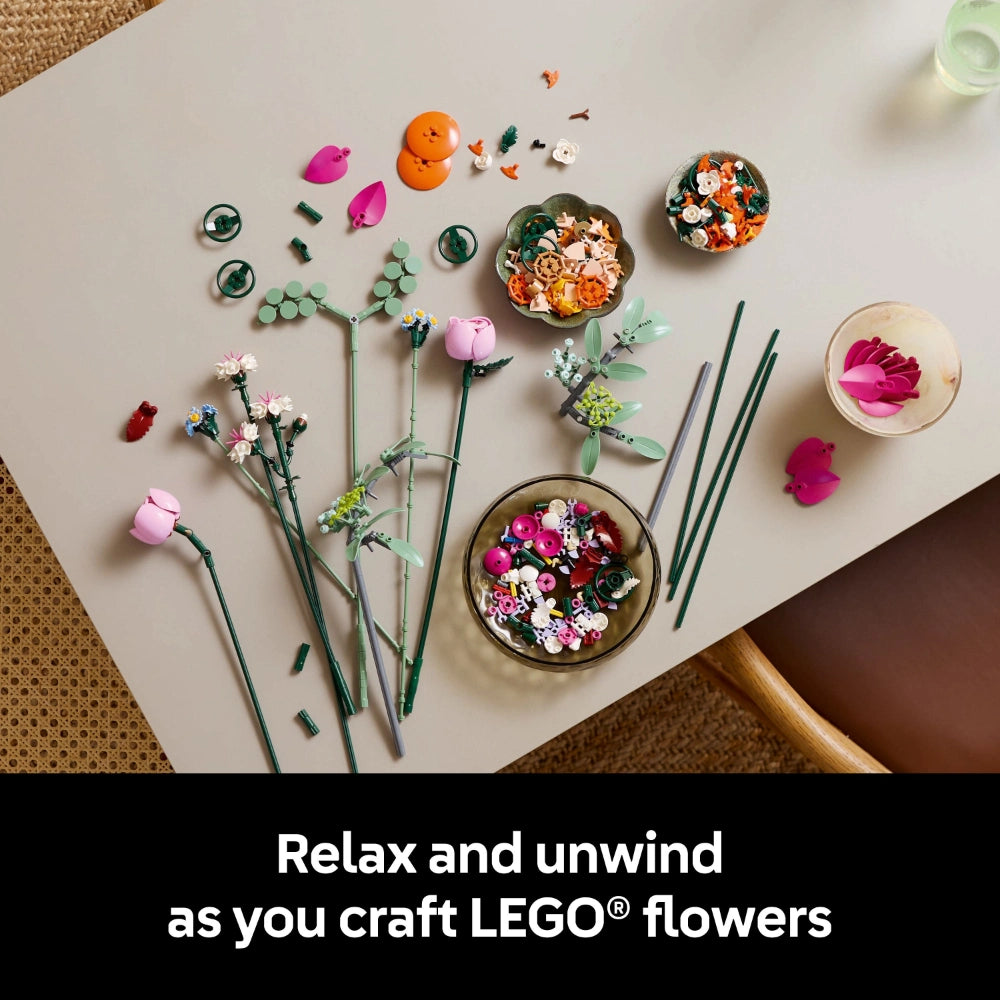 LEGO flower building set display showing colorful assembled blooms in pink, orange, and white with green stems arranged on a white surface, surrounded by individual LEGO pieces and components in various colors including magenta petals, orange flowers, and green foliage elements
